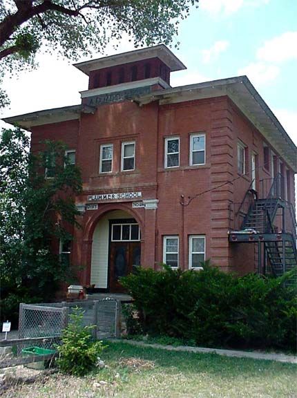 Red brick historic building with arched entrance and exterior fire escape, partly hidden by trees and shrubs.