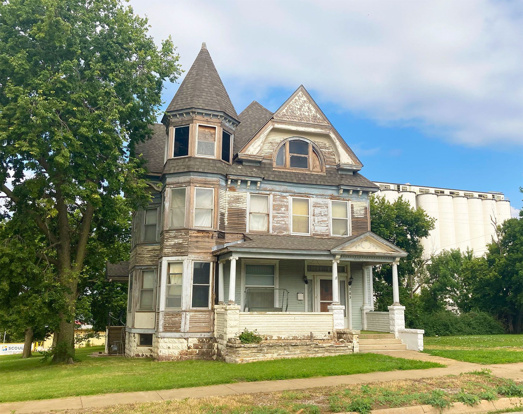 Dilapidated Victorian house with a turret and porch, surrounded by trees under a blue sky