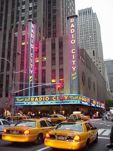 Radio City Music Hall marquee in Manhattan with yellow taxis passing by at dusk.