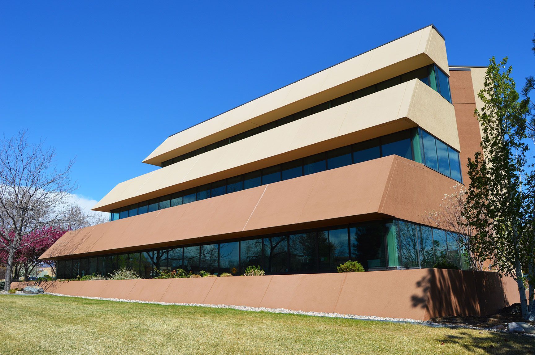 Modern office building with tan and brown tiers, blue windows, and a grassy lawn under a clear sky