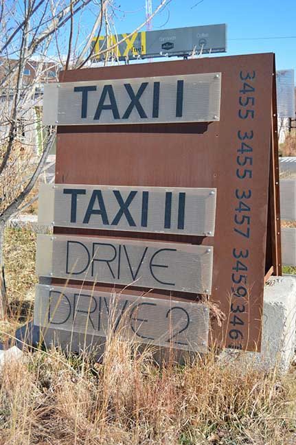 Weathered roadside sign reading “TAXI” and “DRIVE” in a dry, grassy area.