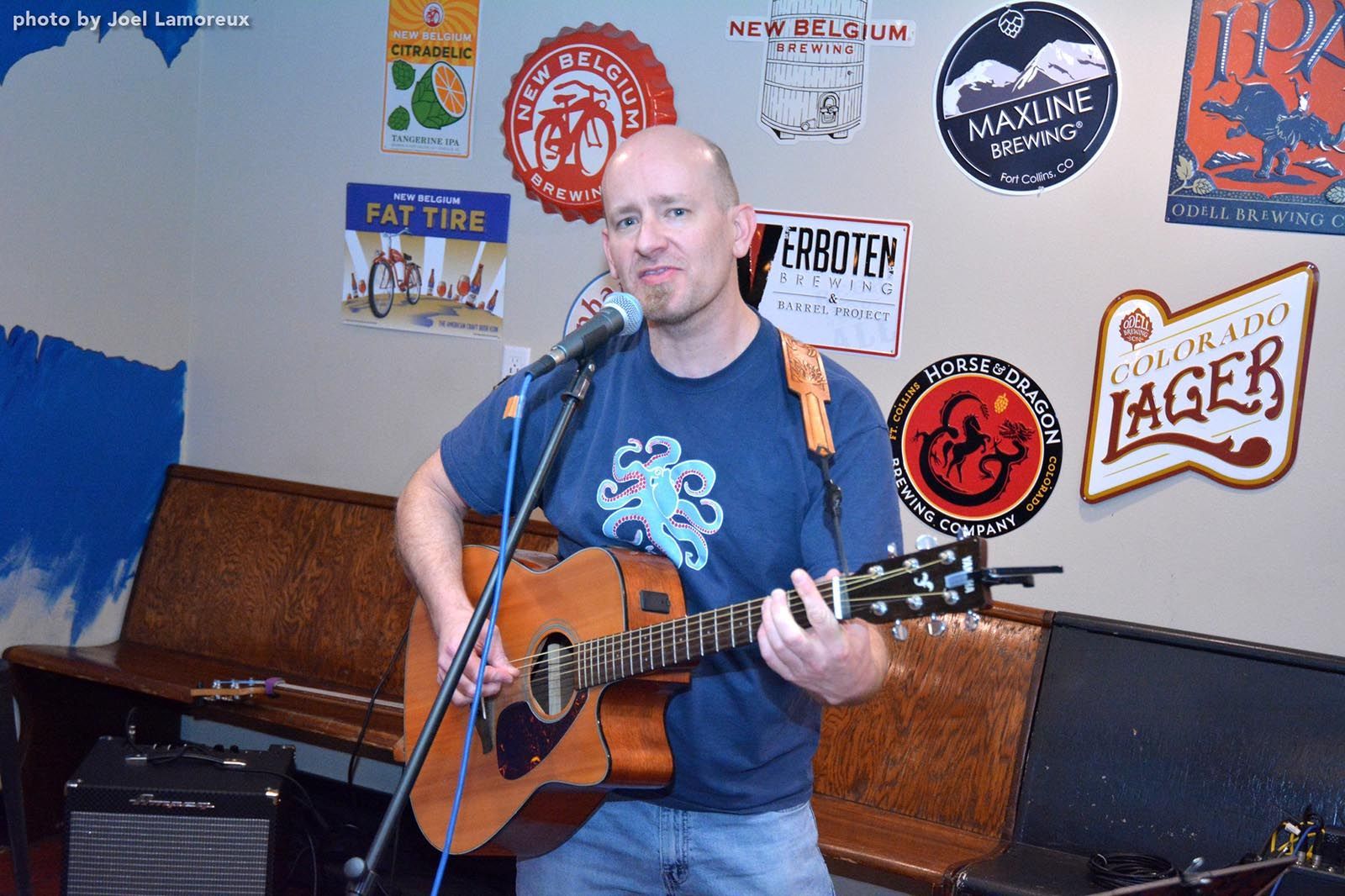 Man singing and playing acoustic guitar onstage in front of colorful beer signs