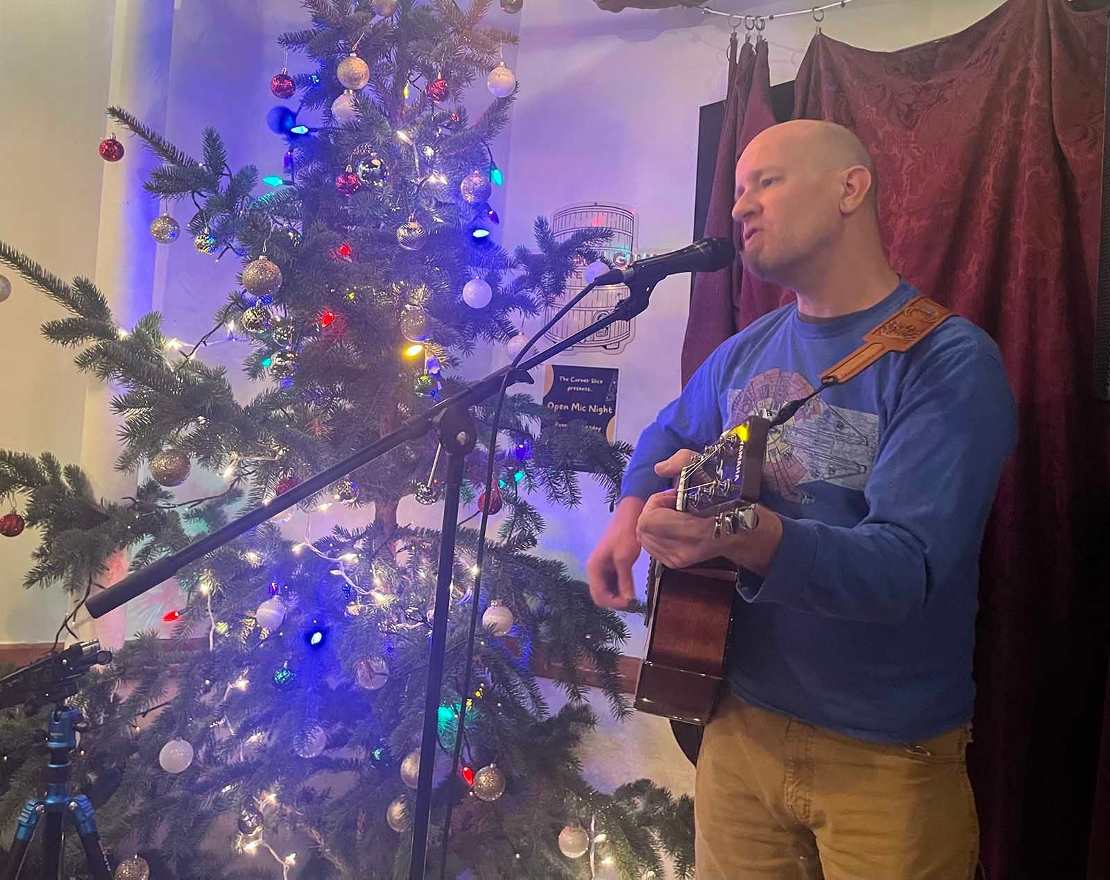 Musician singing and playing guitar beside a lit Christmas tree indoors