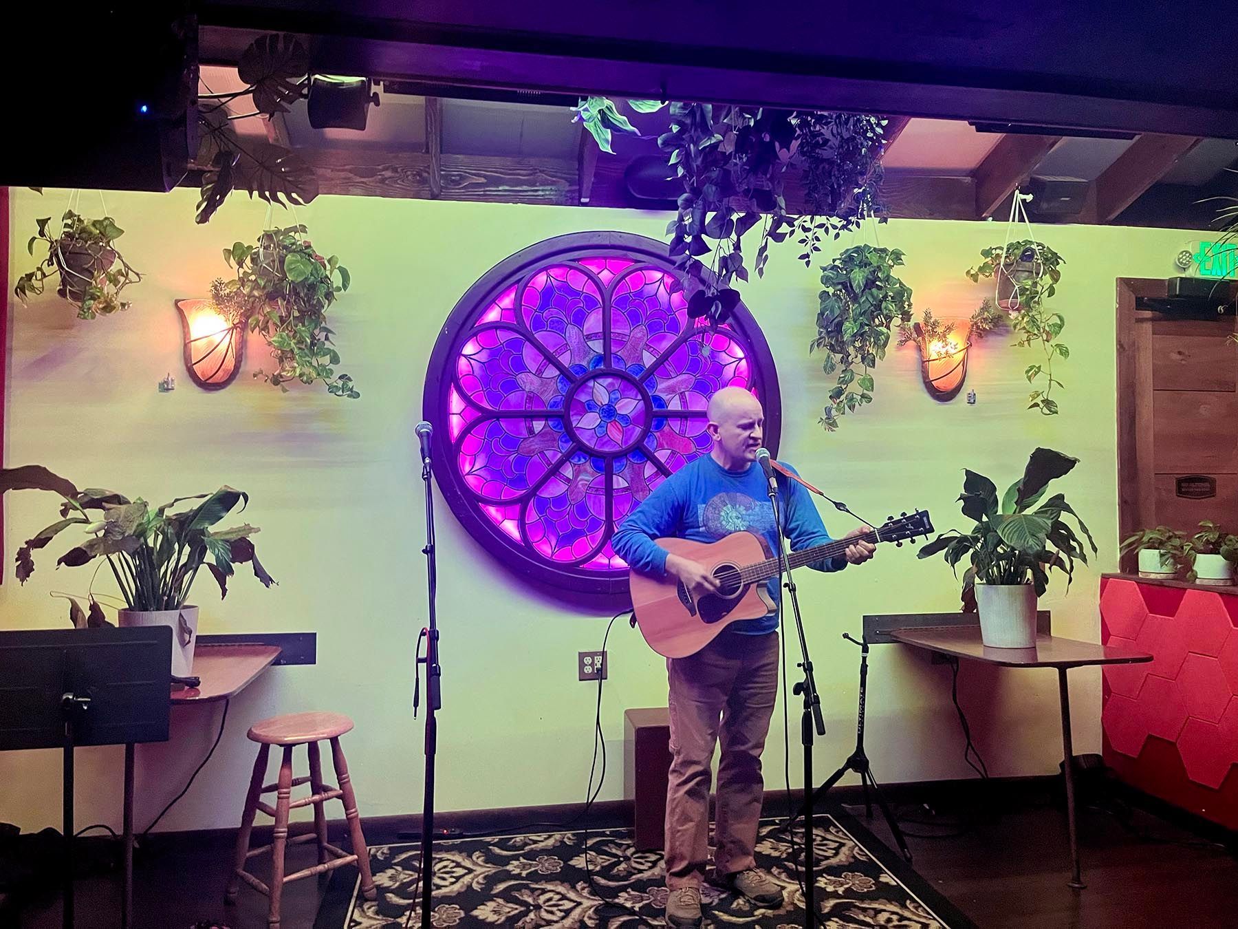 Musician playing guitar on a small stage with purple neon window, plants, and warm wall lights