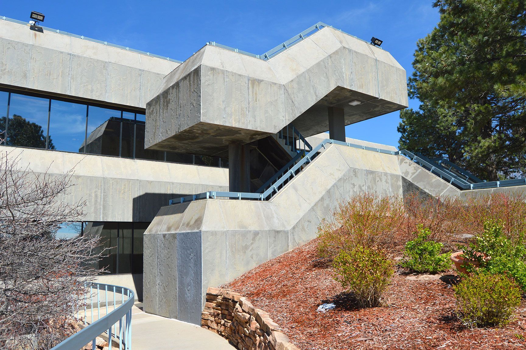 Concrete stairway and elevated walkway beside a modern building, with blue sky and shrubs.
