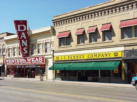 Historic storefronts on a sunny street, including D.J. Penney Company and a western wear shop.