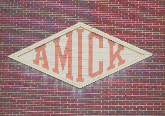 White diamond sign with orange “AMICK” letters on a red brick wall.