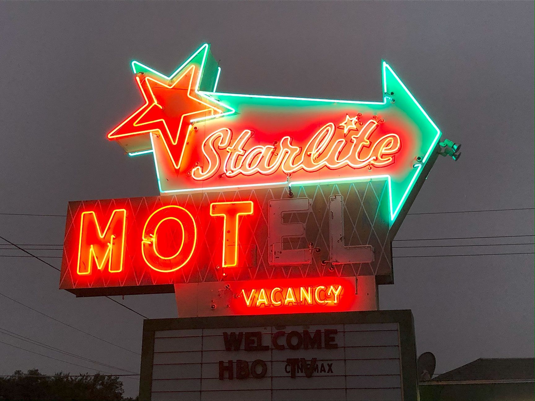 Neon Starlite Motel sign at dusk, glowing red and green with a welcome sign below.