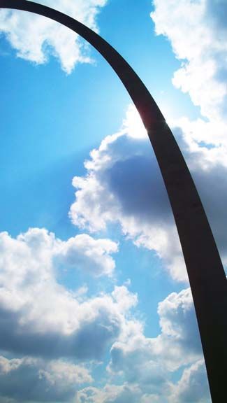 Gateway Arch silhouetted against a bright blue sky with clouds