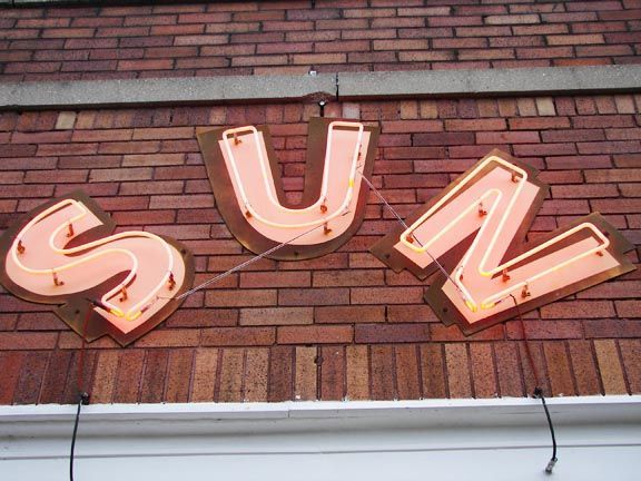 Neon letters spelling “SUN” mounted on a brick wall.