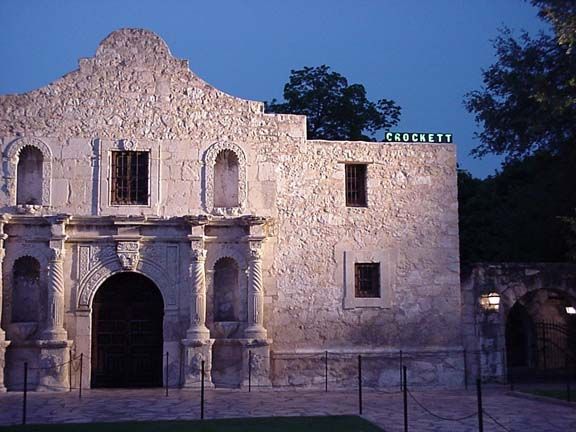 Old stone mission facade at dusk with arched doorway and windows, lit by purple evening sky