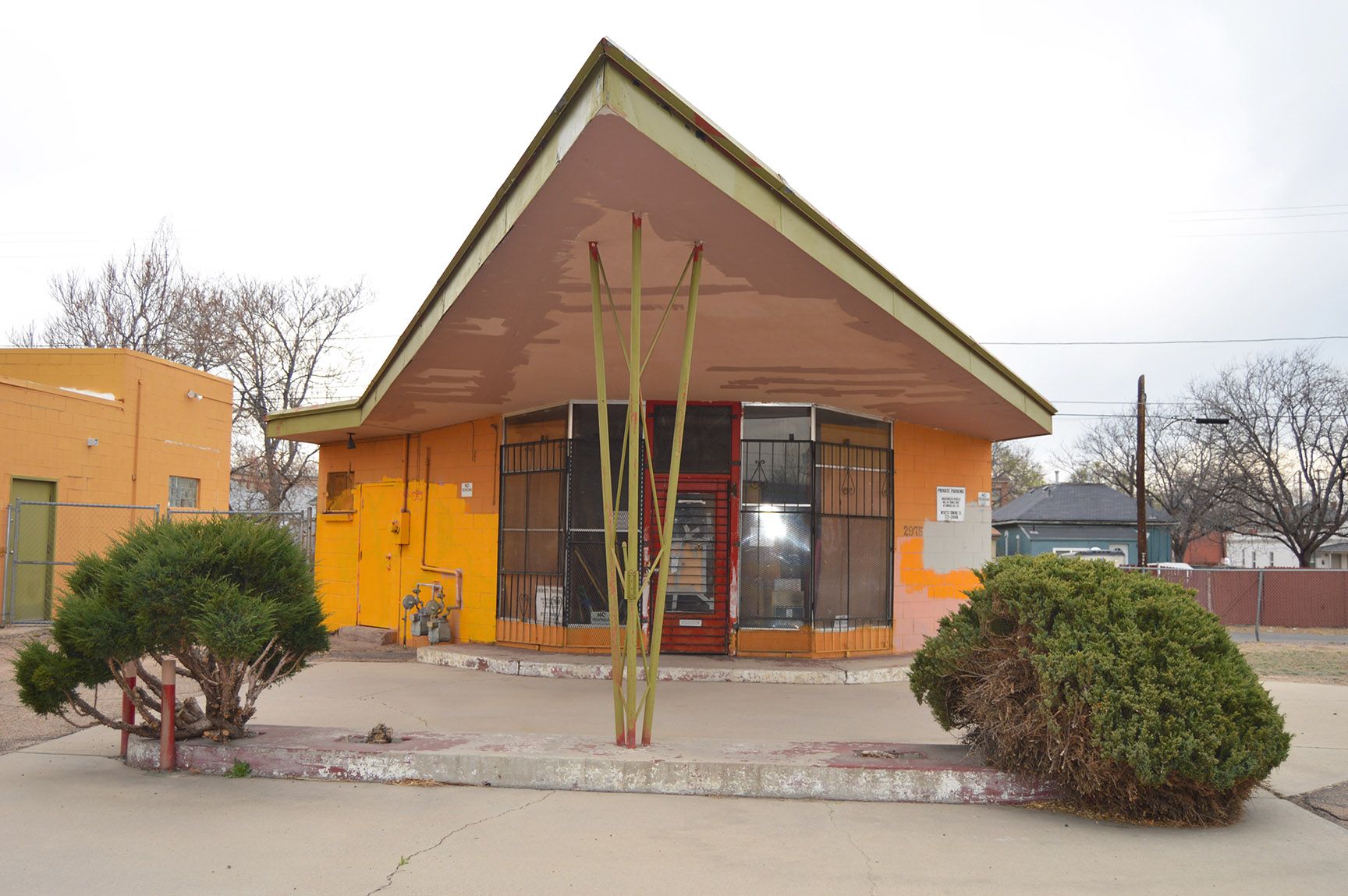 Small yellow building with a peaked roof, glass entrance, and shrubs in front under an overcast sky