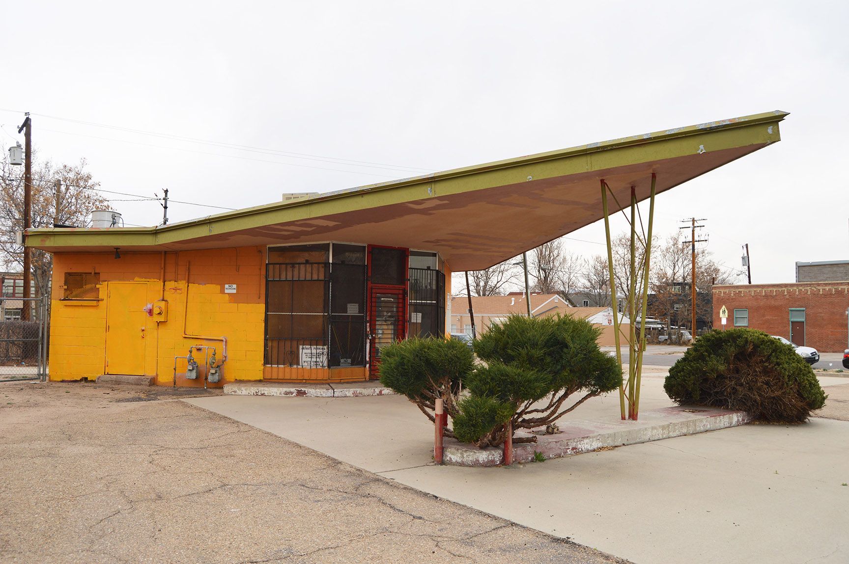 Small orange building with a slanted roof and shrubs outside on a gravel lot