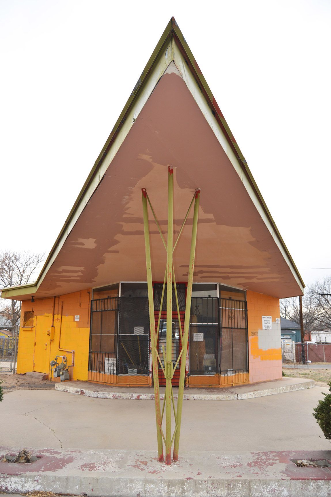 Small orange building with a steep triangular roof and a tall abstract sculpture in front.