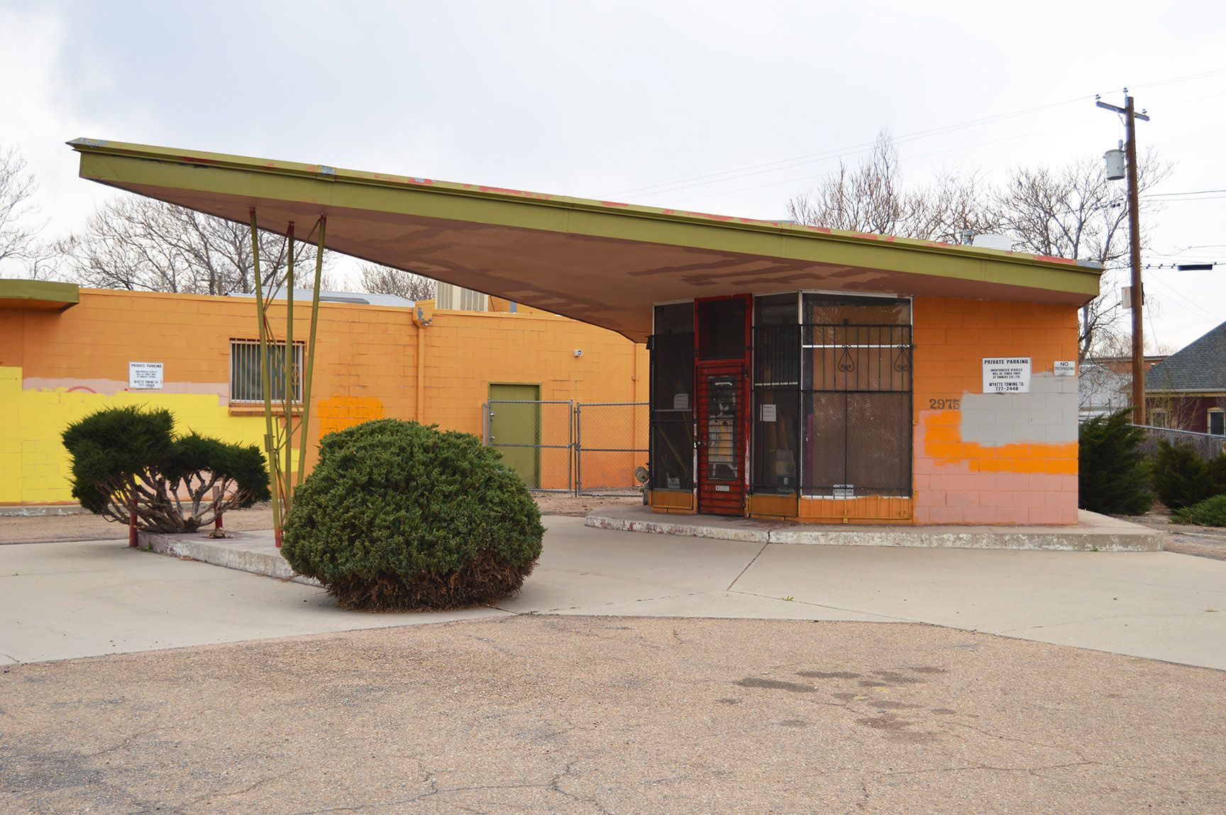 Orange building with a slanted roof, front gate, and small bushes in a gravel lot