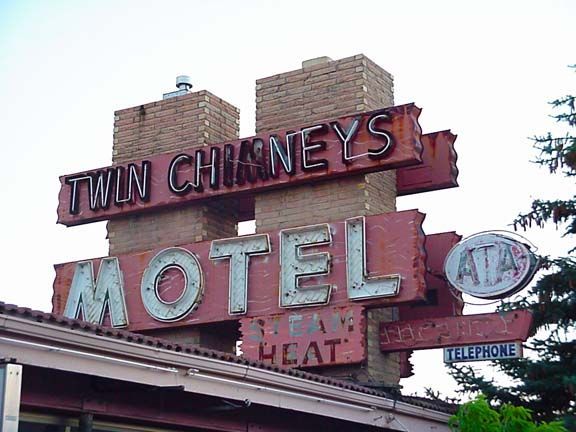 Vintage roadside motel sign reading “Twin Chimneys Motel” with brick chimneys and a telephone icon.
