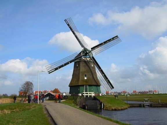 Large windmill by a canal on a cloudy day, with people and red-roofed houses in the distance