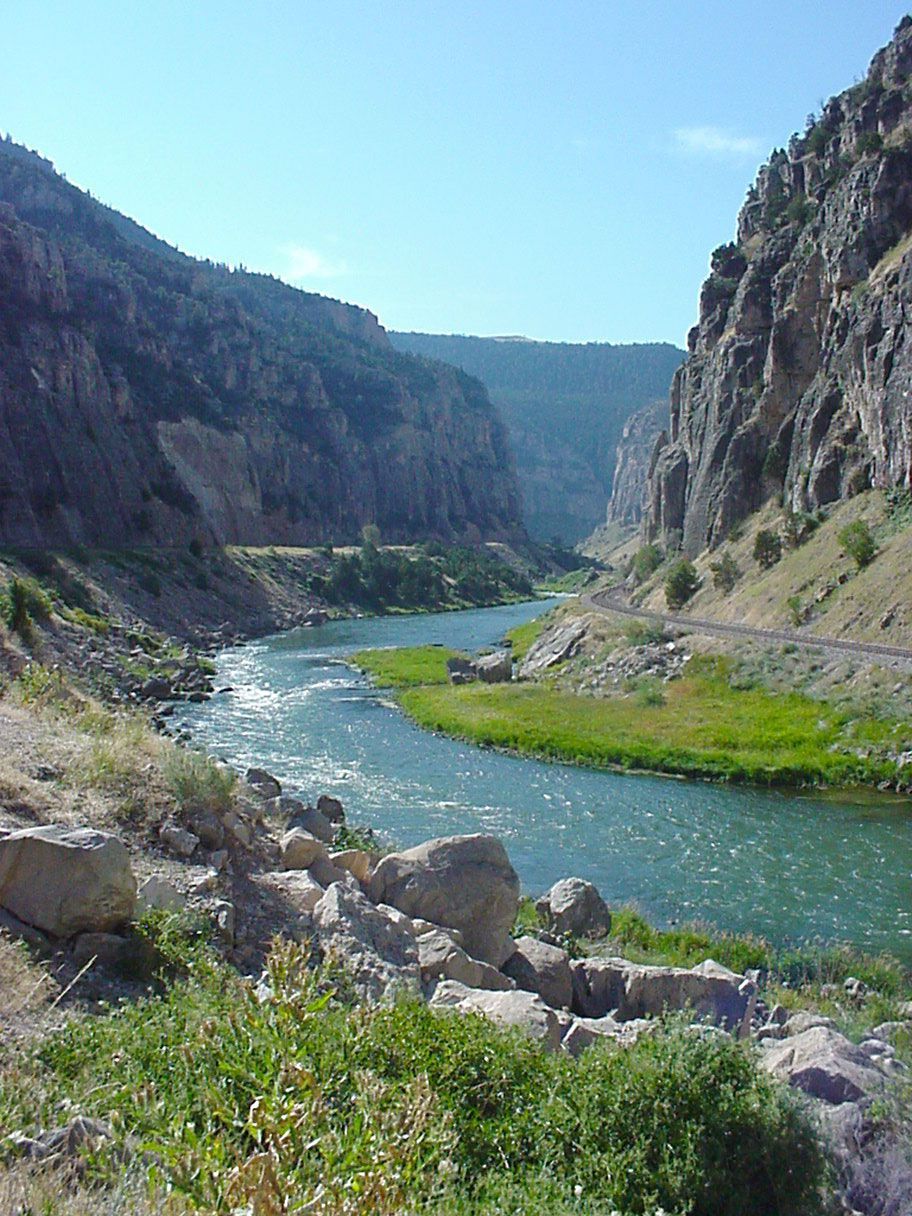Rocky river canyon with blue water, green banks, and steep cliffs under a clear sky, Wind River Canyon, Wyoming