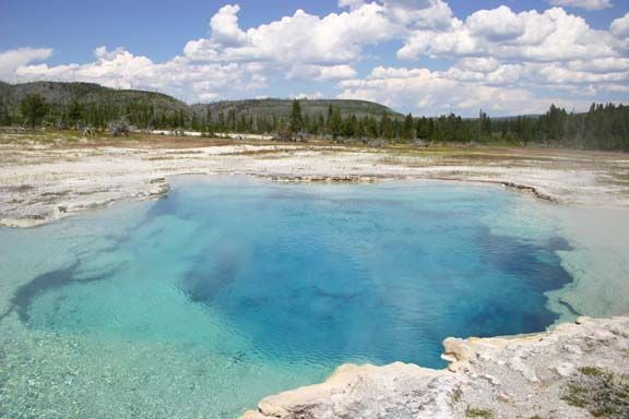 Turquoise hot spring pool surrounded by white mineral deposits and forest under a cloudy sky