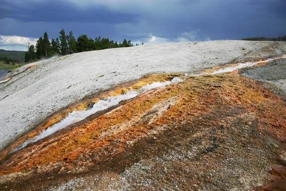 Colorful mineral-stained hillside beneath a dark cloudy sky, with sparse trees in the distance.