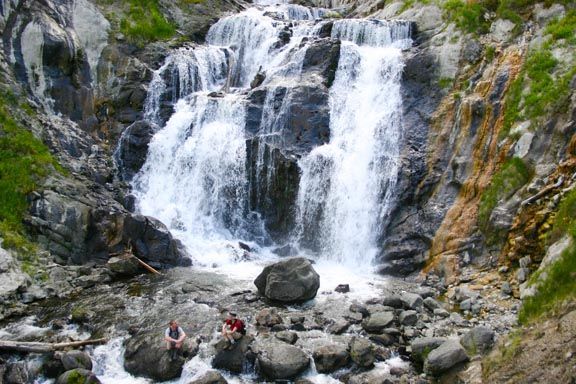 Waterfall cascading over rocky cliffs into a pool, with hikers on boulders below, Mystic Falls, Yellowstone National Park, Wyoming