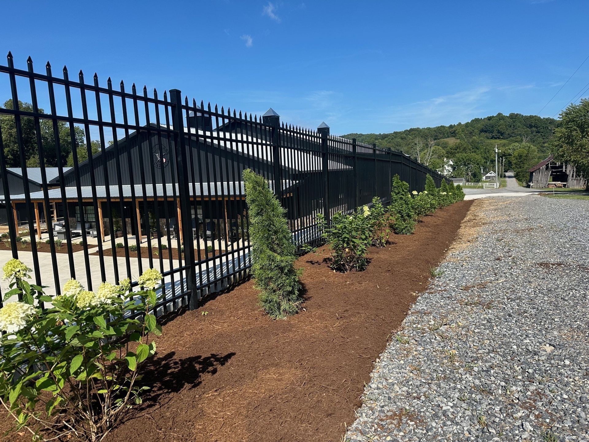 Black metal fence with landscaped border along a gravel driveway; building in the background.