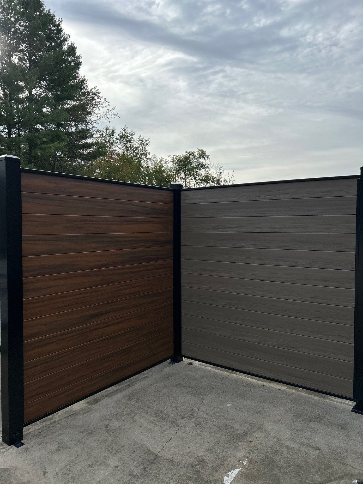 Two-tone composite fence, brown and gray horizontal planks, black posts, on concrete. Cloudy sky background.