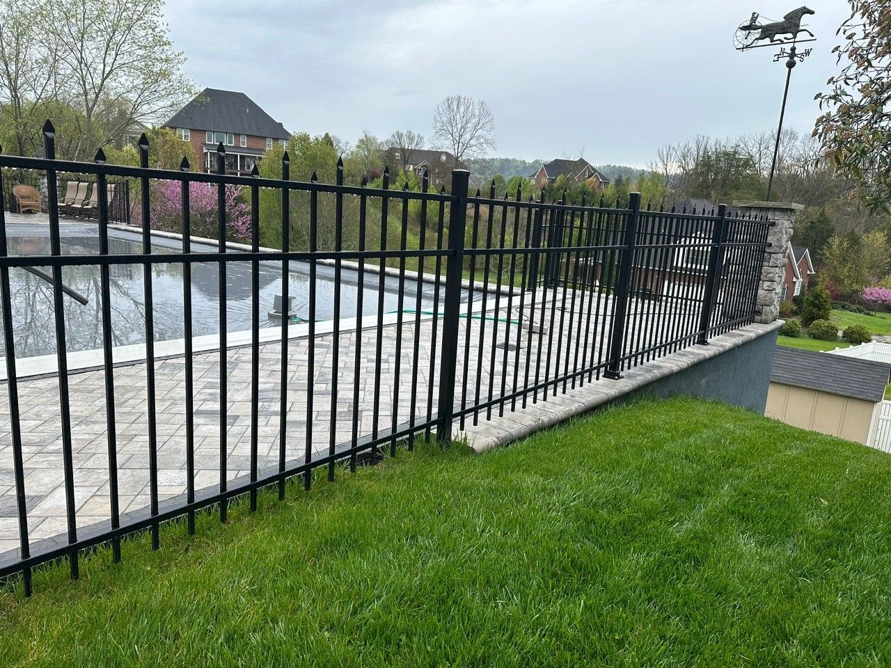 Black metal fence along a stone-lined pool deck with green lawn. Horse weathervane in the distance.