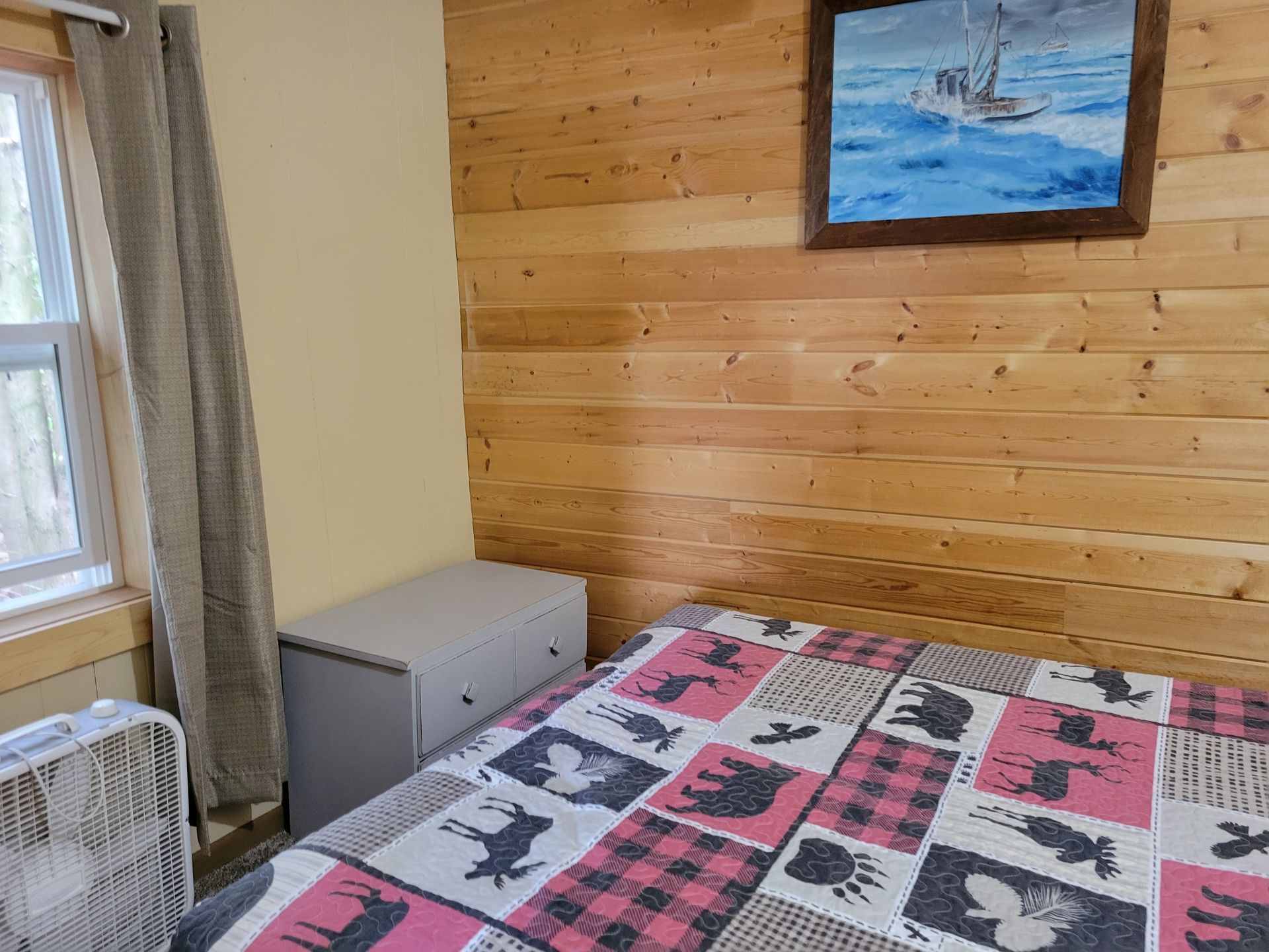 A bedroom with a bed , dresser , fan and a painting on the wall.
