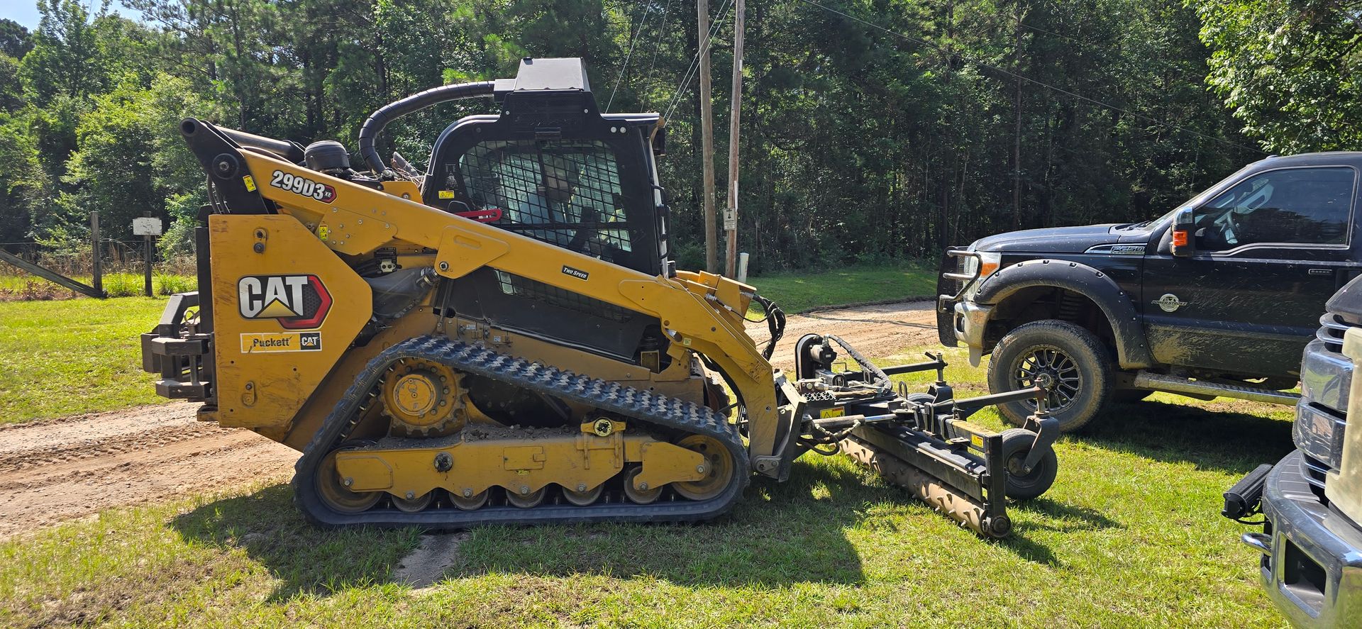 A yellow CAT skid steer with a grading attachment on a grassy area next to a black truck and trees.