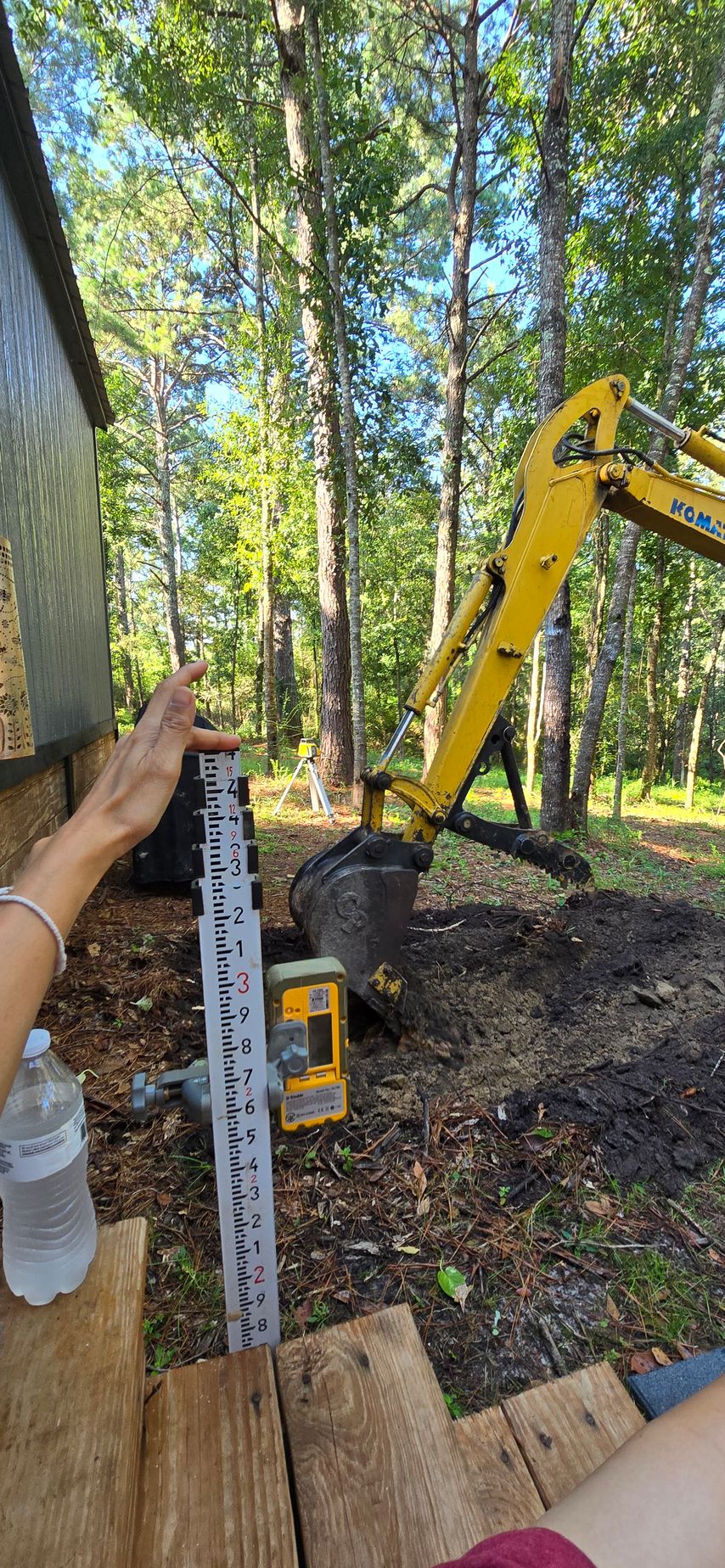 Person using a ruler next to an excavator in a wooded area.