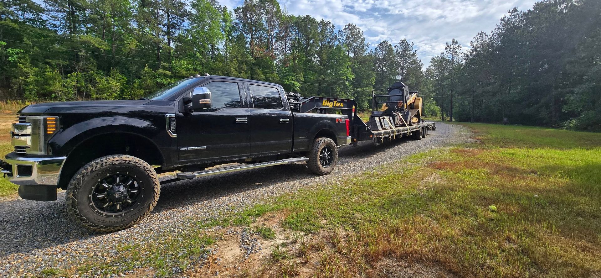 Black truck towing equipment on a gravel road, surrounded by trees and green grass.
