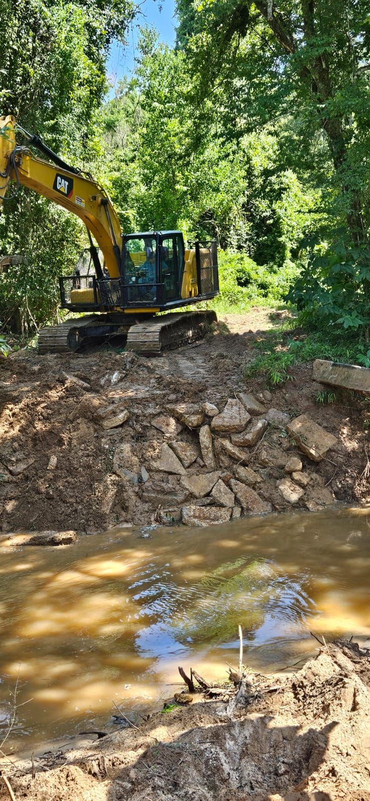 An excavator working by a stream in a wooded area.