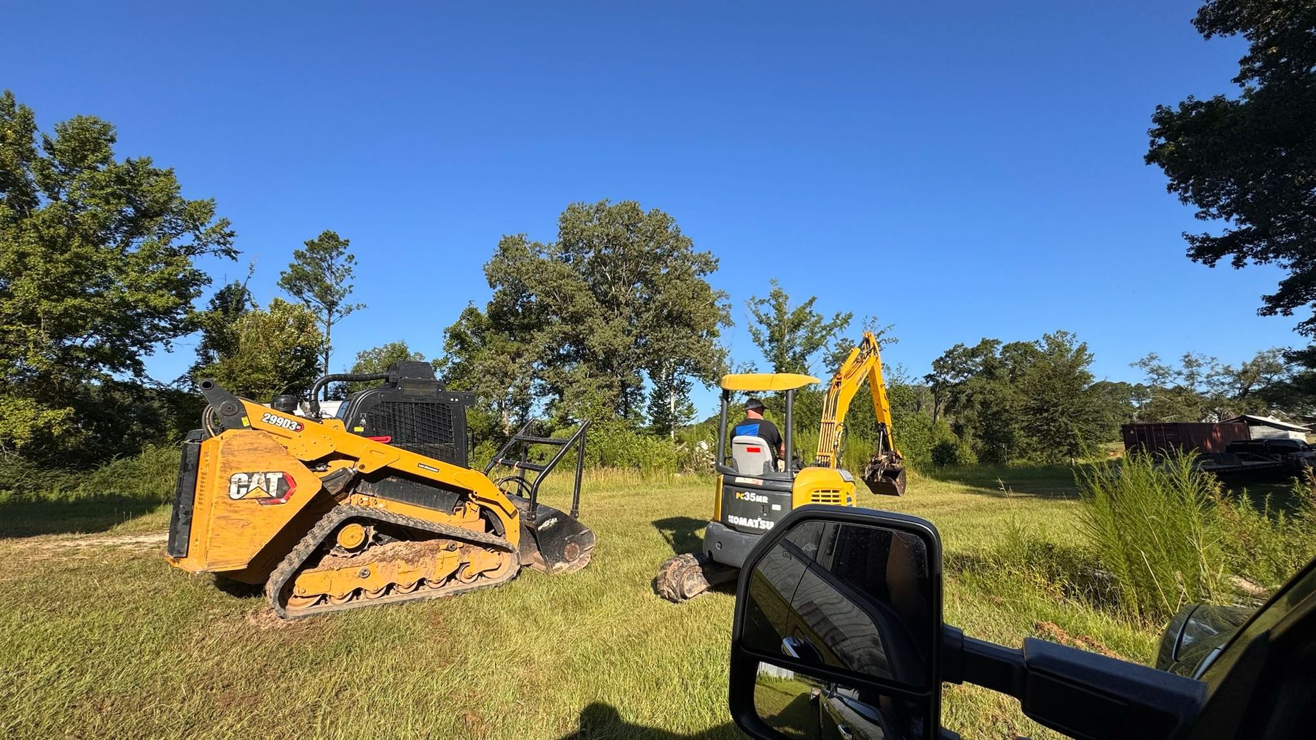Yellow CAT skid steer and excavator on a grassy field under a blue sky.