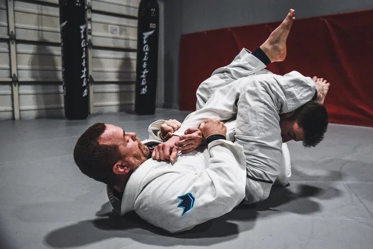 Two people practicing jiu-jitsu on a mat, one in a white gi pinned on his back.
