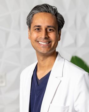 Smiling doctor in a white coat, blue scrubs, standing in a clinic setting.