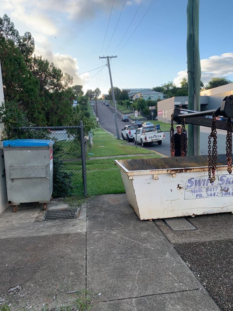 A sloping street with vehicles, a bin, and a skip bin. Trees on the left and power lines overhead. — Swift Skips in Gympie, QLD