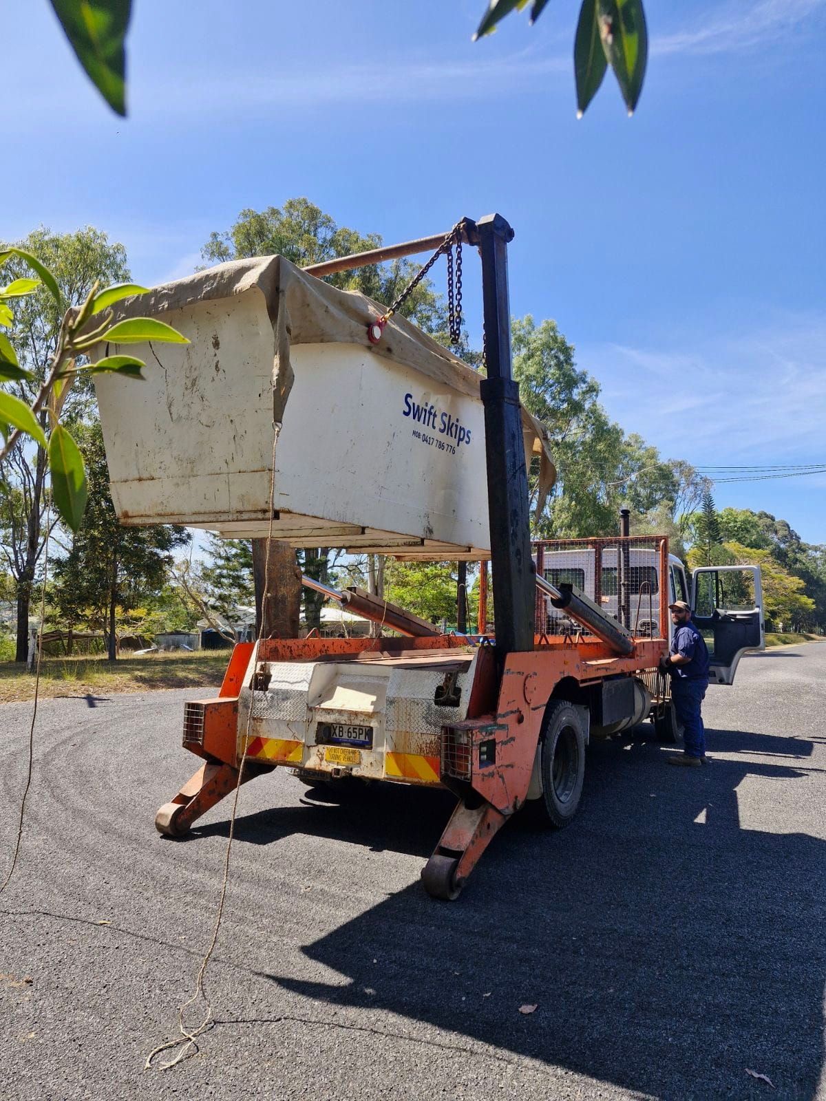 Orange truck with a bin being lifted by a crane. A person stands nearby. Sunny day. — Swift Skips in Mary Valley, QLD