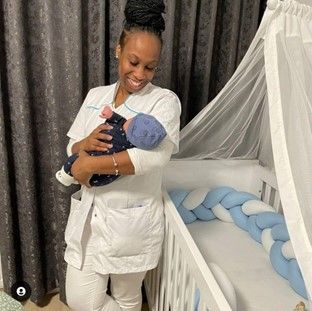 Vrouw in wit uniform met een baby op haar arm, lachend naast een wieg met een blauw-witte rand.