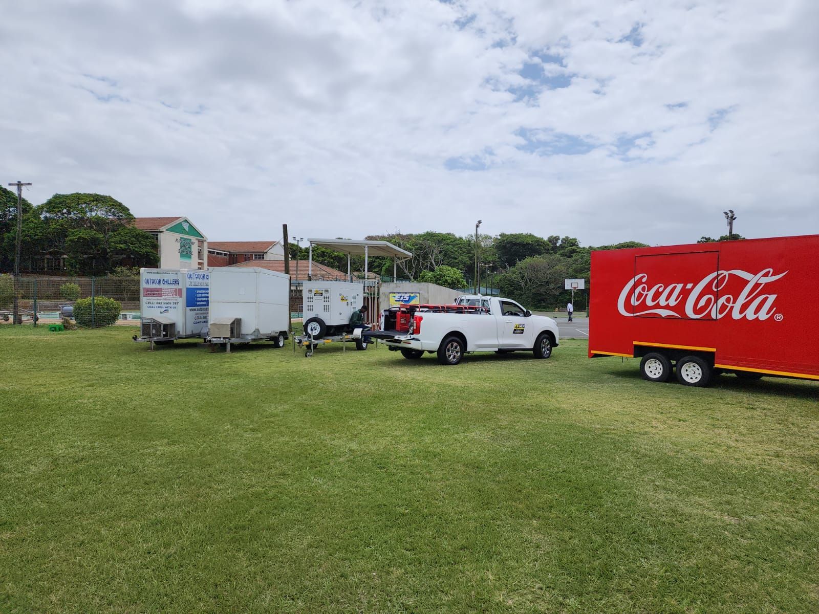 A coca cola trailer is parked in a grassy field next to a white truck.