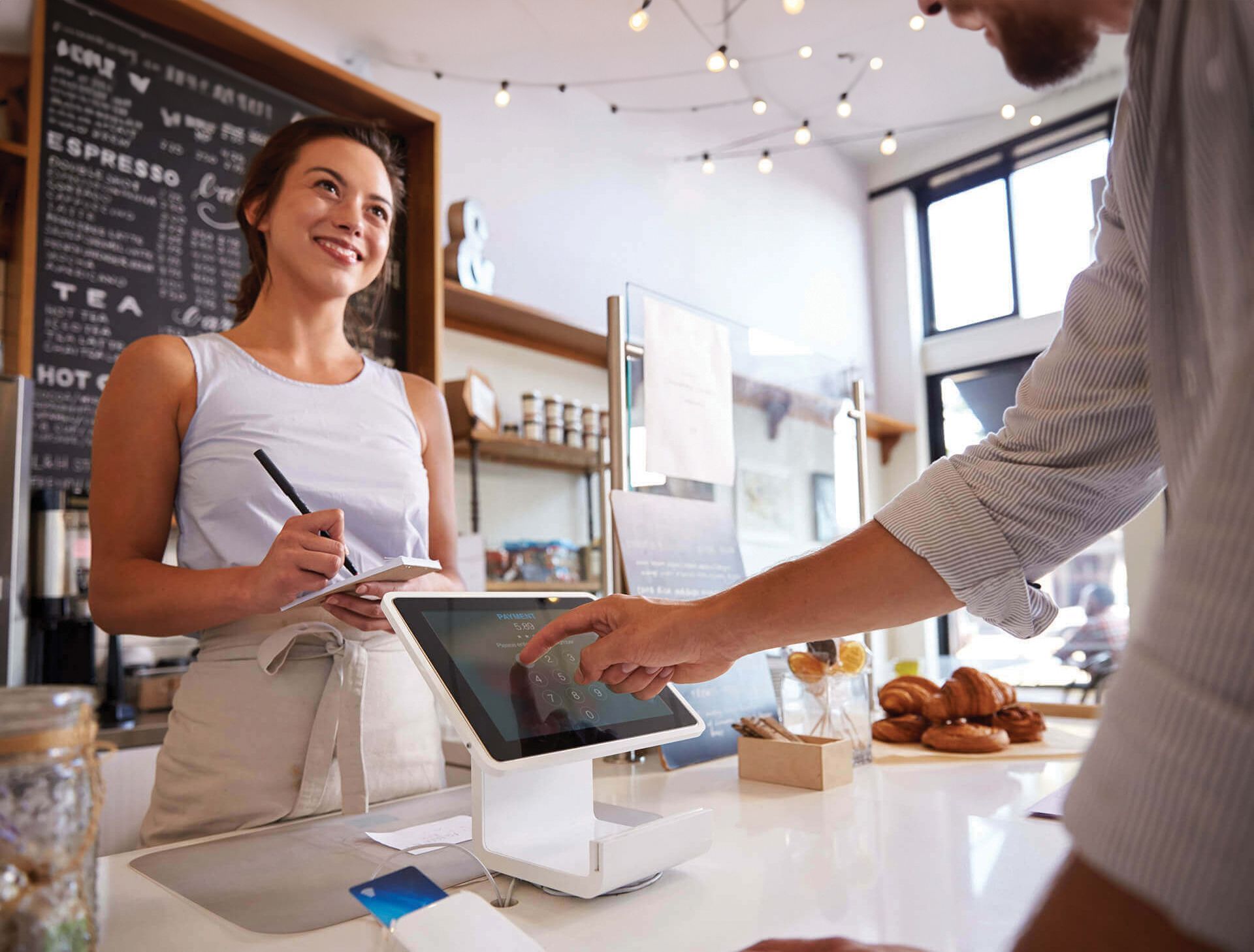 Barista using a tablet at a bright café counter while a customer pays nearby