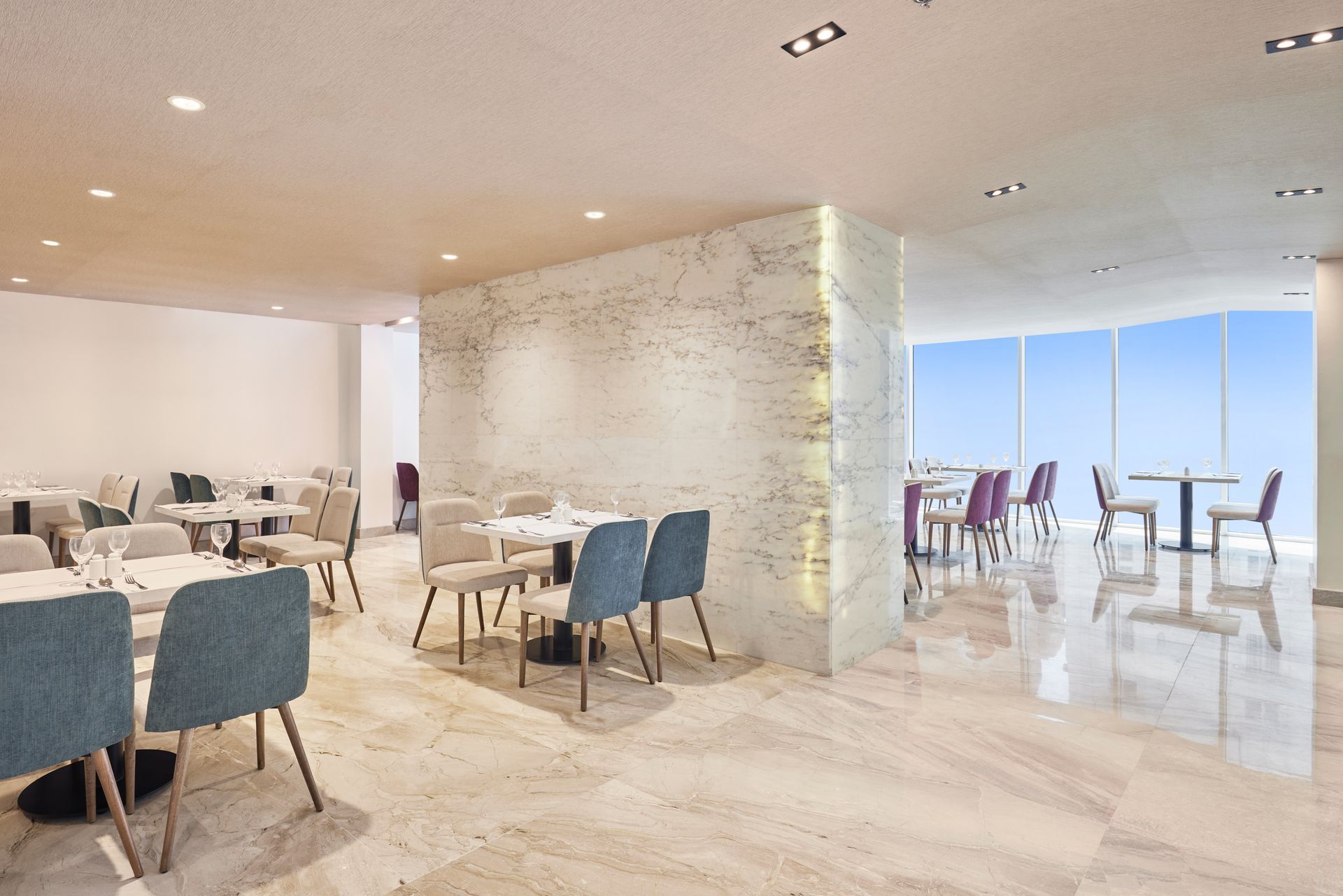 Bright modern restaurant with marble floors, blue chairs, and tables by large windows overlooking the sea