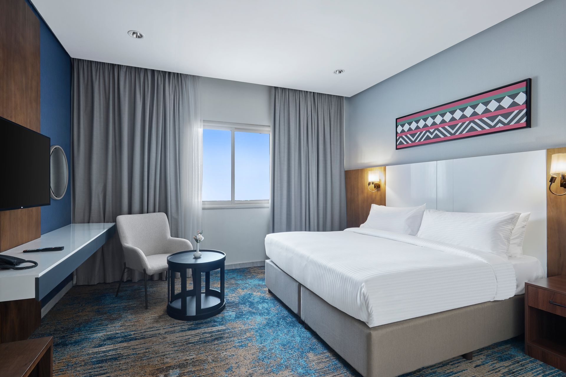 Modern hotel room with queen bed, window, gray curtains, chair, and blue patterned carpet