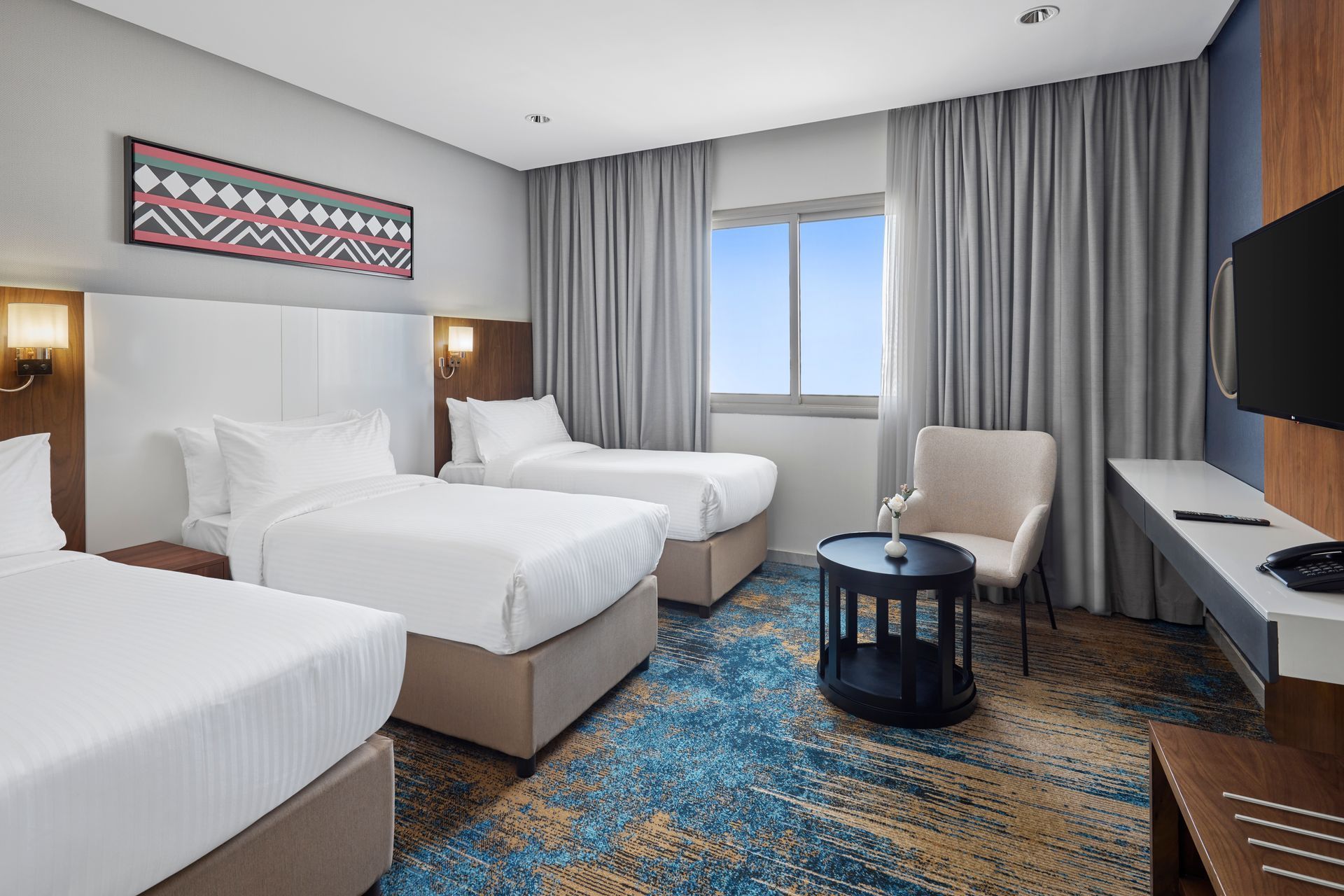Modern hotel room with three beds, window, chair, TV, and patterned blue carpet.