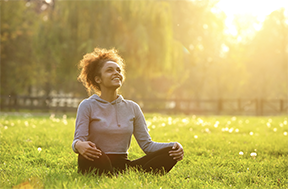 A woman is sitting in a lotus position in the grass in a park.