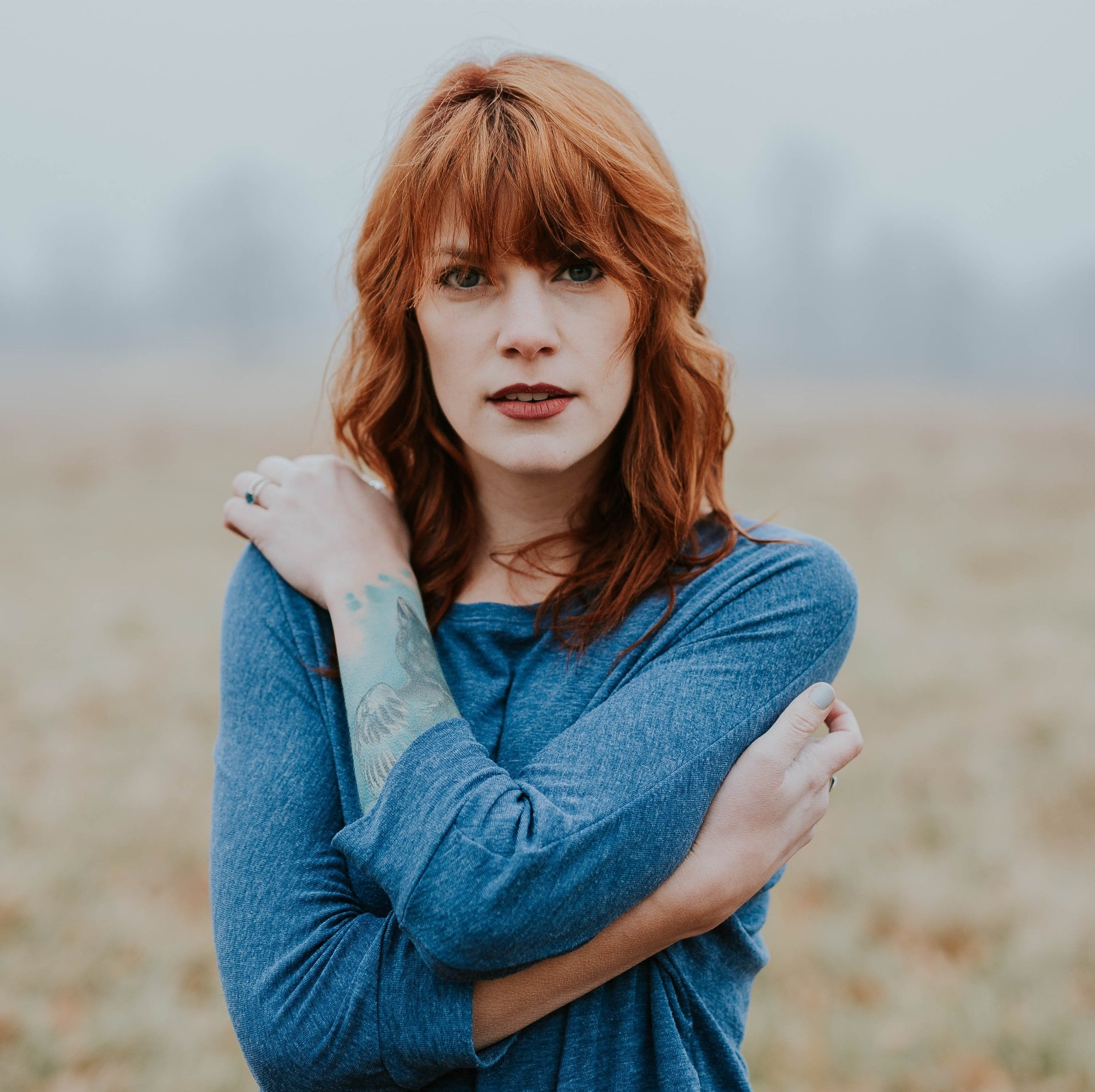 A woman with red hair is standing in a field with her arms crossed.