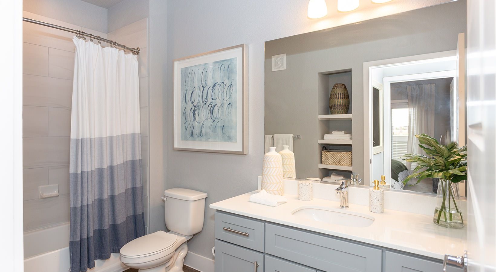 Modern apartment bathroom with double vanity, large mirror, toilet, shower with striped curtain, and shelves.