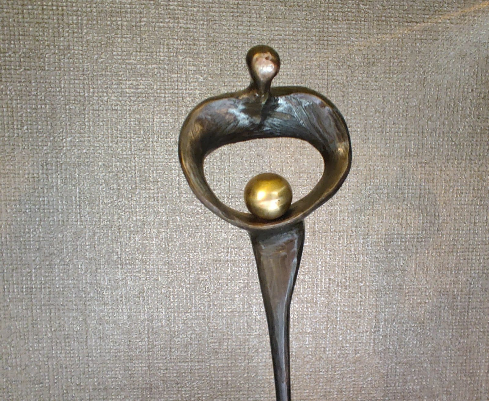 Bronzen sculptuur van een figuur die een gouden bol vasthoudt, geplaatst tegen een gestructureerde, grijze achtergrond.