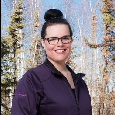 women with glasses on A woman wearing glasses and a purple jacket is smiling in front of trees.