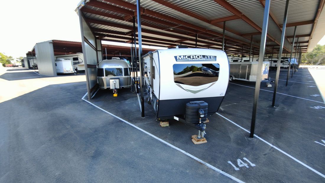 RVs parked under a metal awning in a storage lot; asphalt ground with numbered parking spots.